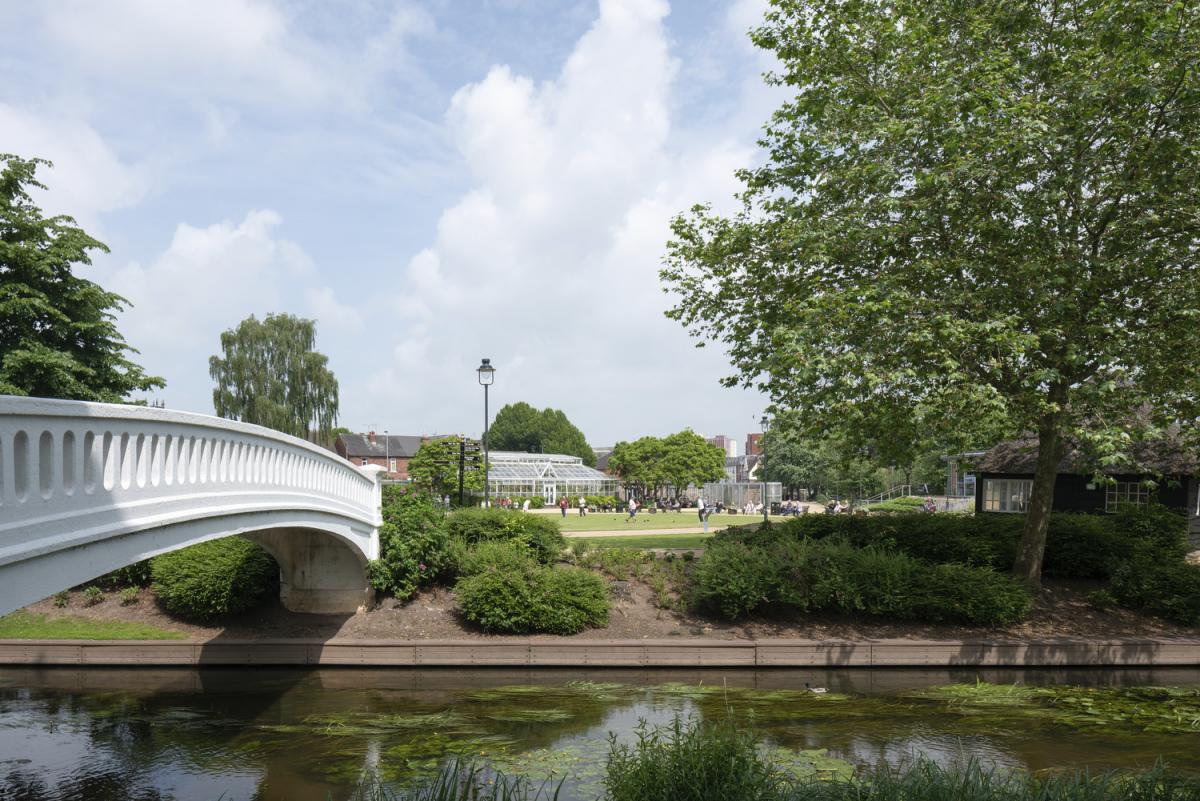 a view across a river in a park to people playing bowls, with a white bridge crossing to the left and a large tree to the right