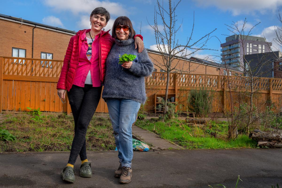 two ladies, one wearing a bright pink jacket and the other a blue jumper,have their arms around each smiling at the camera. They are in an city centre allotment.
