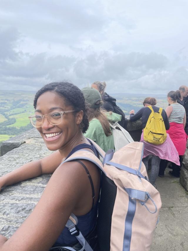 a lady with glasses and a pink backpack smiles at the camera, she is with a group of people walking, they are all looking out at the landscape.