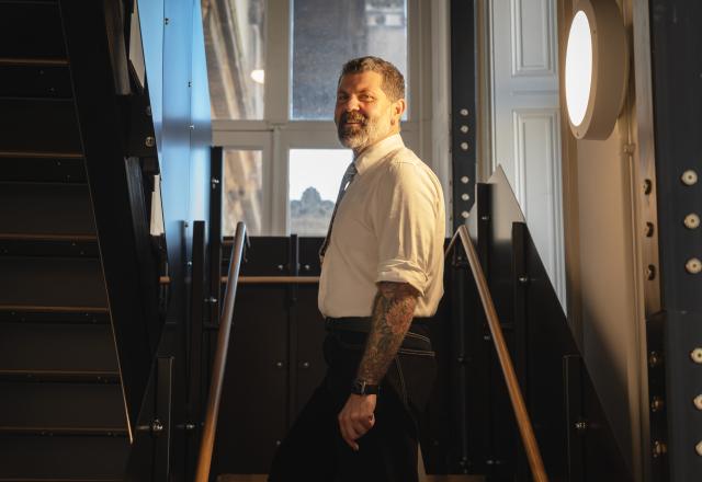 Man in a white shirt with rolled up sleeves, standing side on in a room with a window behind him.