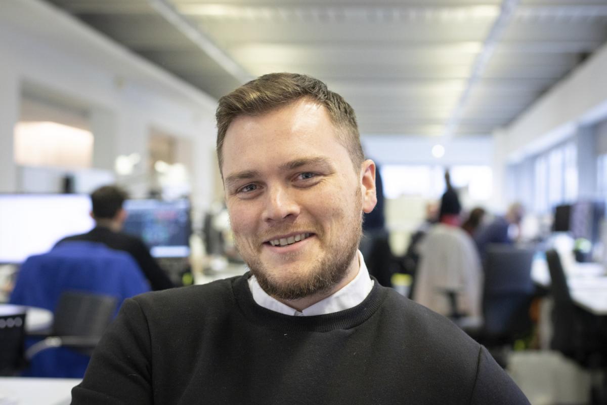 Headshot of Alex Scrimshaw. A man smiling, wearing a black jumper and a white shirt.