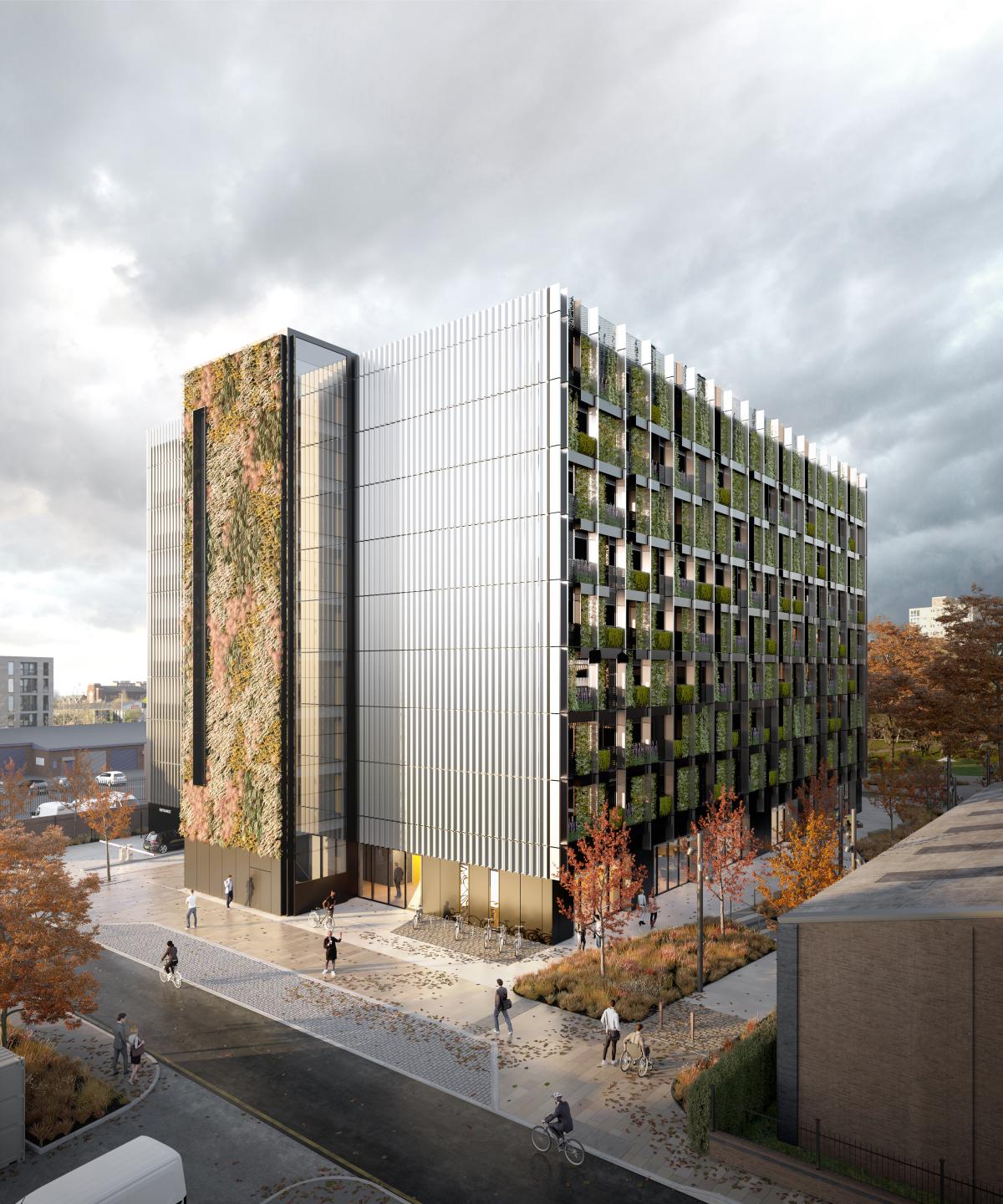 Large building, partly covered by a living planted wall. It is surrounded by young trees and people walking to and from the building.  