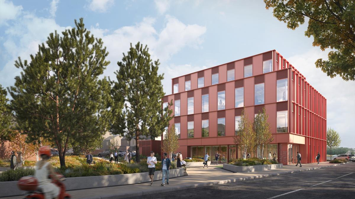 Outside view. Tree lined street. Three storey pink/red building with glazing. The ground floor has a large glazed front. Infront of the building is a planted green space.