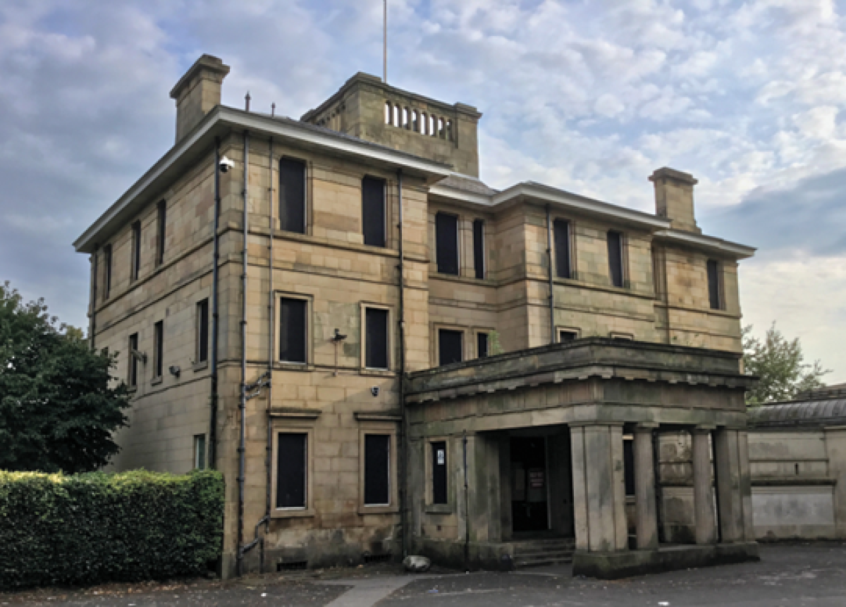 Large grand Victorian mansion building in pale/yellow stone. It's three storeys with an extended porch, probably for carriages to draw up to. Although the building looks to be in a good condition, the windows are boarded up with black boards.