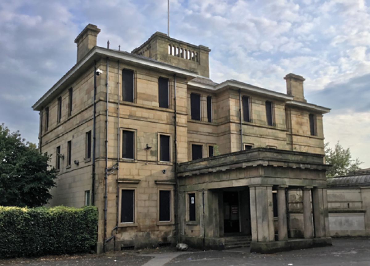 Large grand Victorian mansion building in pale/yellow stone.  It's three storeys with an extended porch, probably for carriages to draw up to.  Although the building looks to be in a good condition, the windows are boarded up with black boards.  