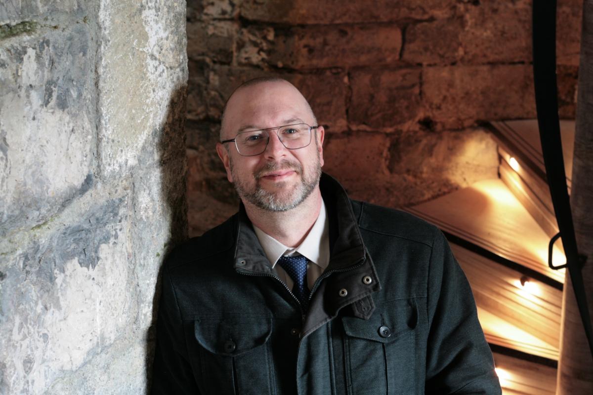 A man in a black jacket stands in front of an enclosed stone and timber spiral staircase at Caernarfon Castle