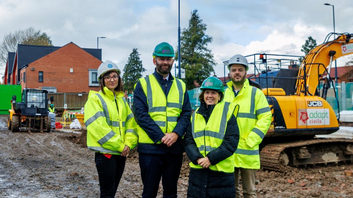 External photograph of a small construction site.  There are four people in the picture, smiling at the camera, wearing high visibility jackets and construction helmets.  