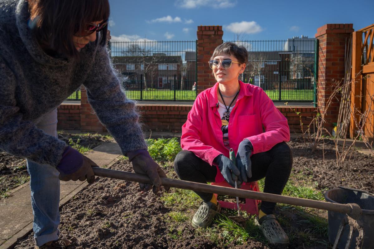 a lady in a pink jacket squats down as a lady in a blue jumper hoes the ground in front of her.