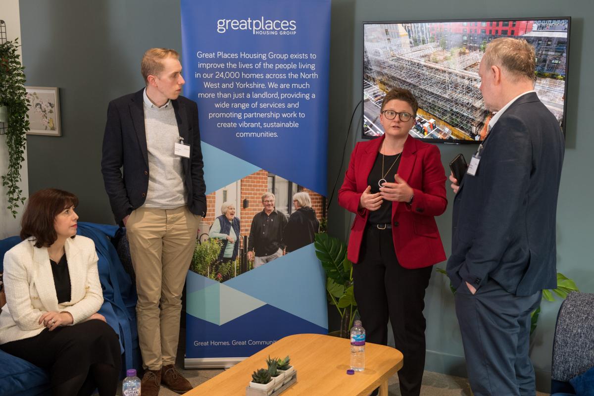 Internal photo of four people. Three people are standing infront of a presentation screen displaying a scaffolded building. Two men stand either side of a woman in a red jacket, Cllr Bev Craig. Another woman is sitting in a chair listening.