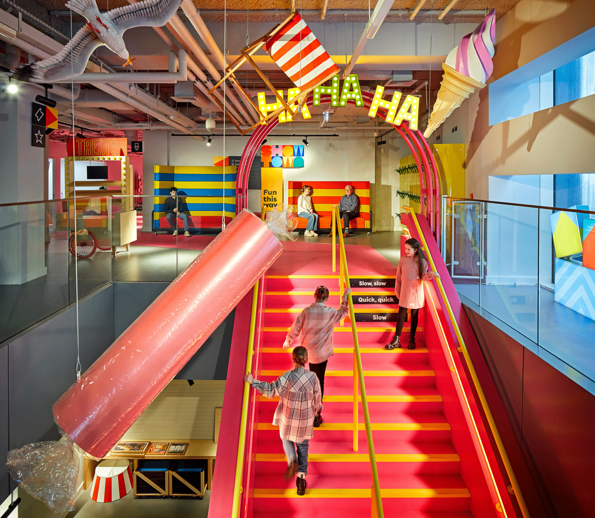 three girls walk up the pink stairs to Showtown in Blackpool. oversized sculptures of rock and deckchairs hang from the ceiling, and ha ha ha is an illuminated sign at the top.