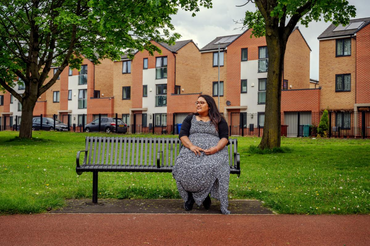 a lady sits on a bench in a park. A row of brick houses sit behind.