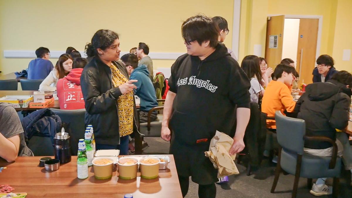 a room full of people a man in a black hooded top is in the foreground talking to a lady in a yellow top and black jacket