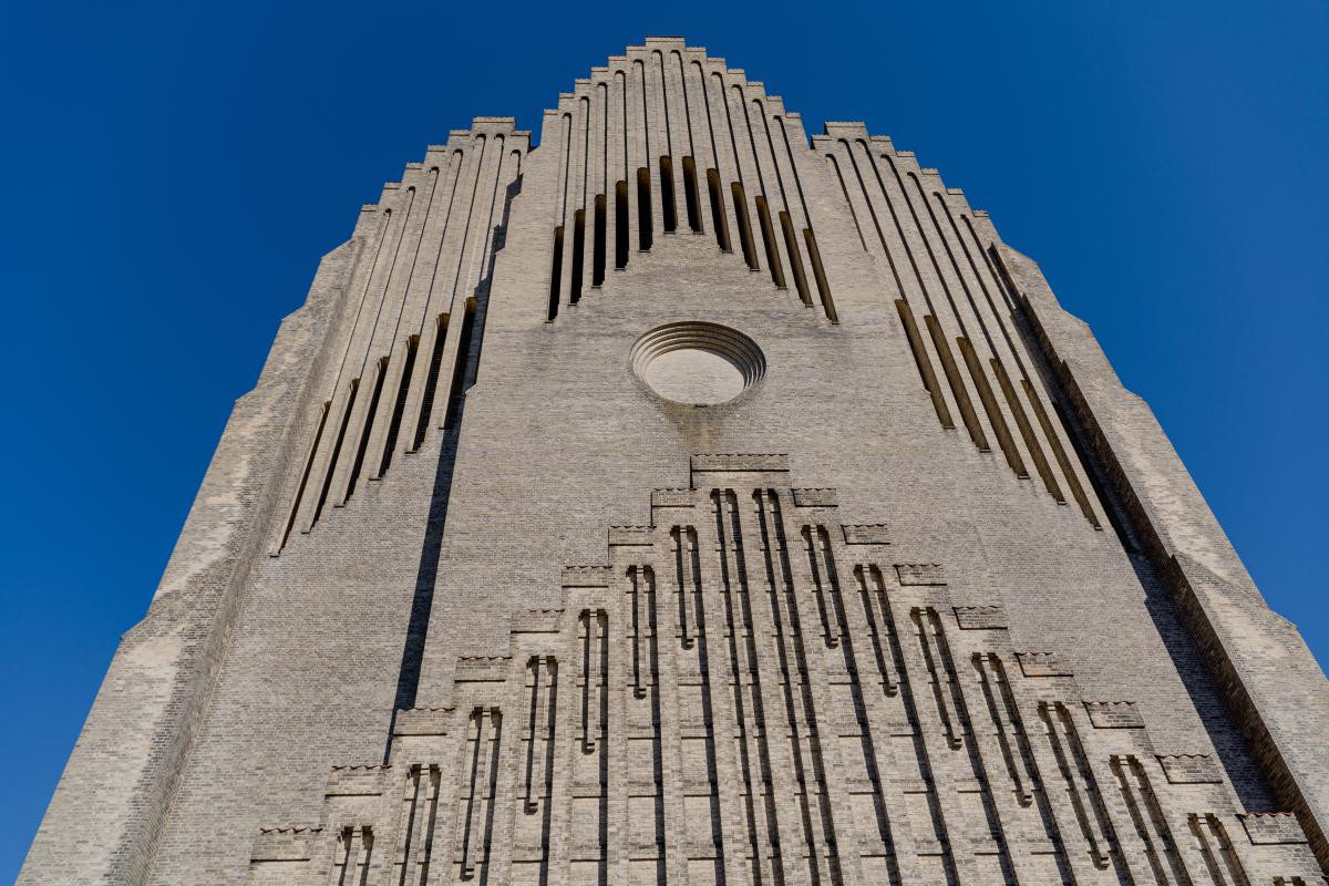 Image taken from the ground looking up at the front facade of the church. It is a very grey concrete like facade which contrasts with the blue sky. There are lines working vertically which form part of a triangle shape which decorate the facade.  There is an indented circle in the