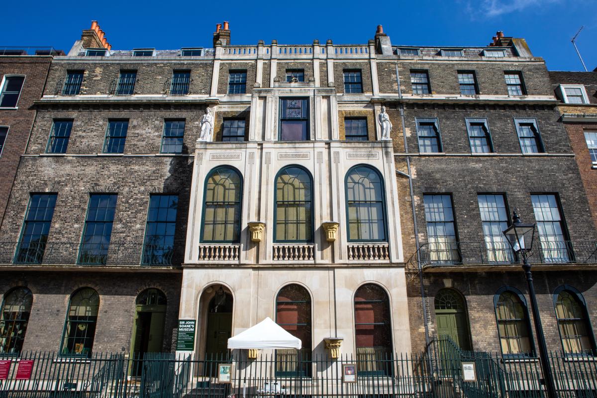 Street view of buildings. Four storeys. The central block is a white colour which has large arch windows at first floor.