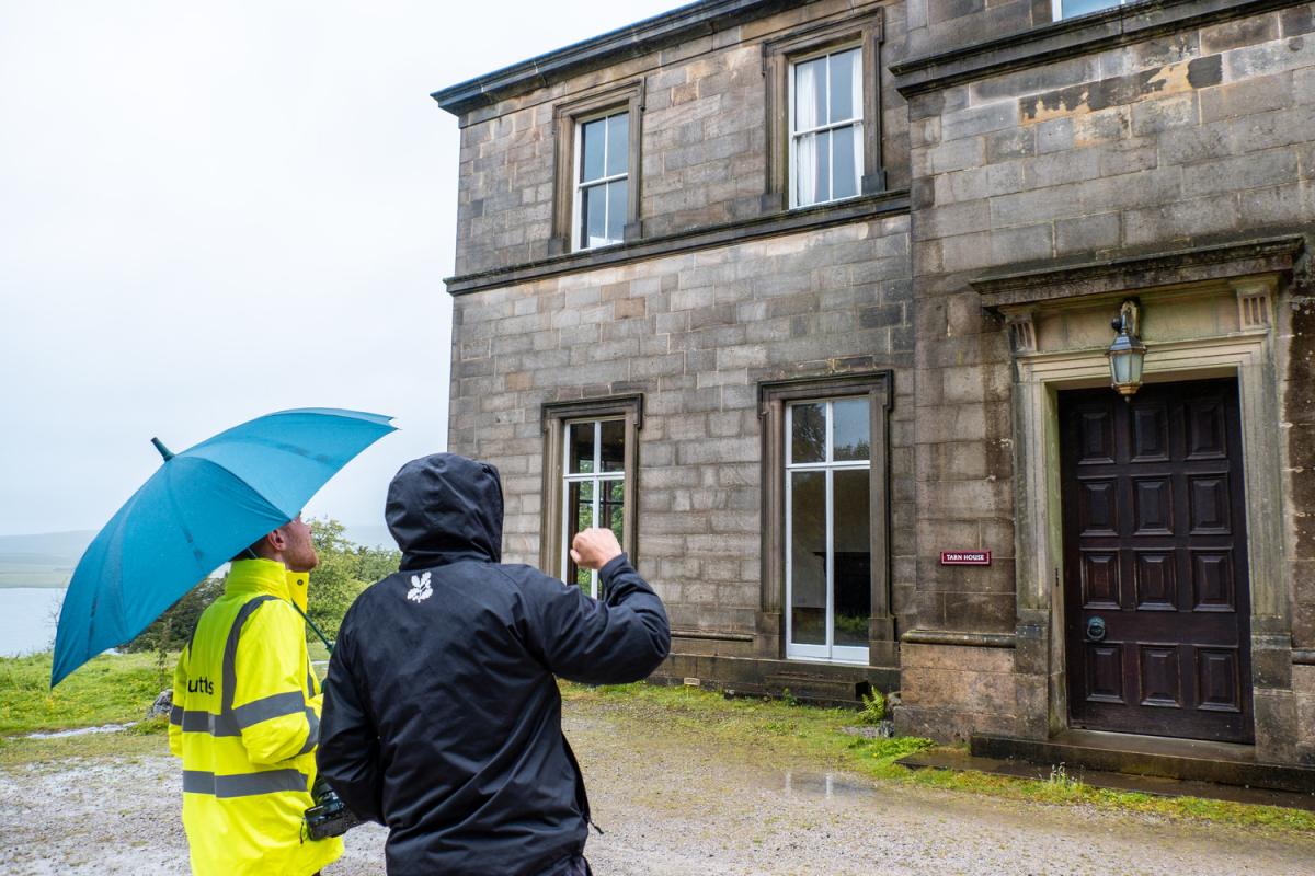 two men, one in a high vis jacket and using a blue umbrella, are looking at the front of a historic stone building