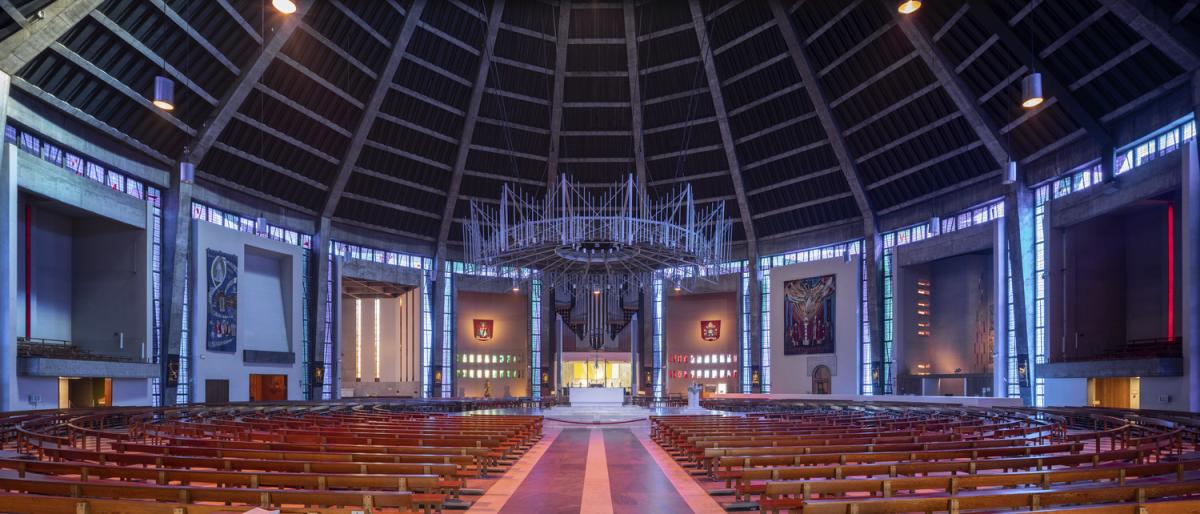 Internal photograph of Liverpool Metropolitan Cathedral. It's a circular building with rich colours of red, purple and yellow. Seating is laid out in a circular fan-style with the pulpit in the middle of the circle.  