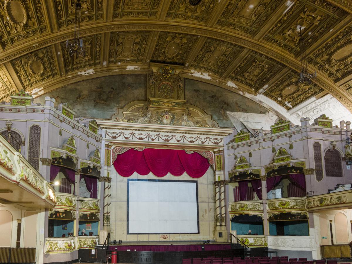 Internal view of a Victorian concert hall. Showing the stage area and stately boxes for guests on either side of the stage. The ceiling is gold yellow colour and very ornate. 