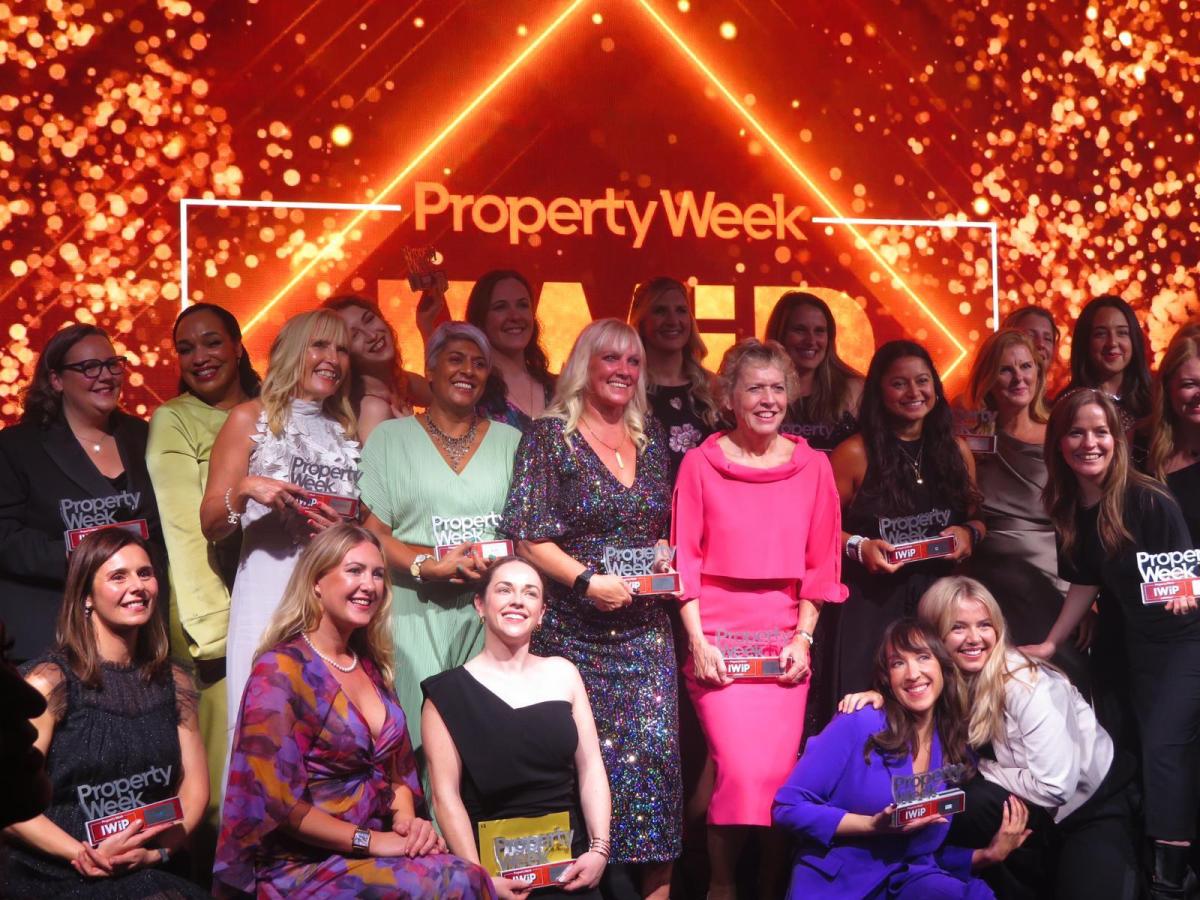 A group of women 'all the winners' stand for their photograph at the end of the awards event. Behind them is a sparkly back drop with Property Week Awards logo in bright colours. 