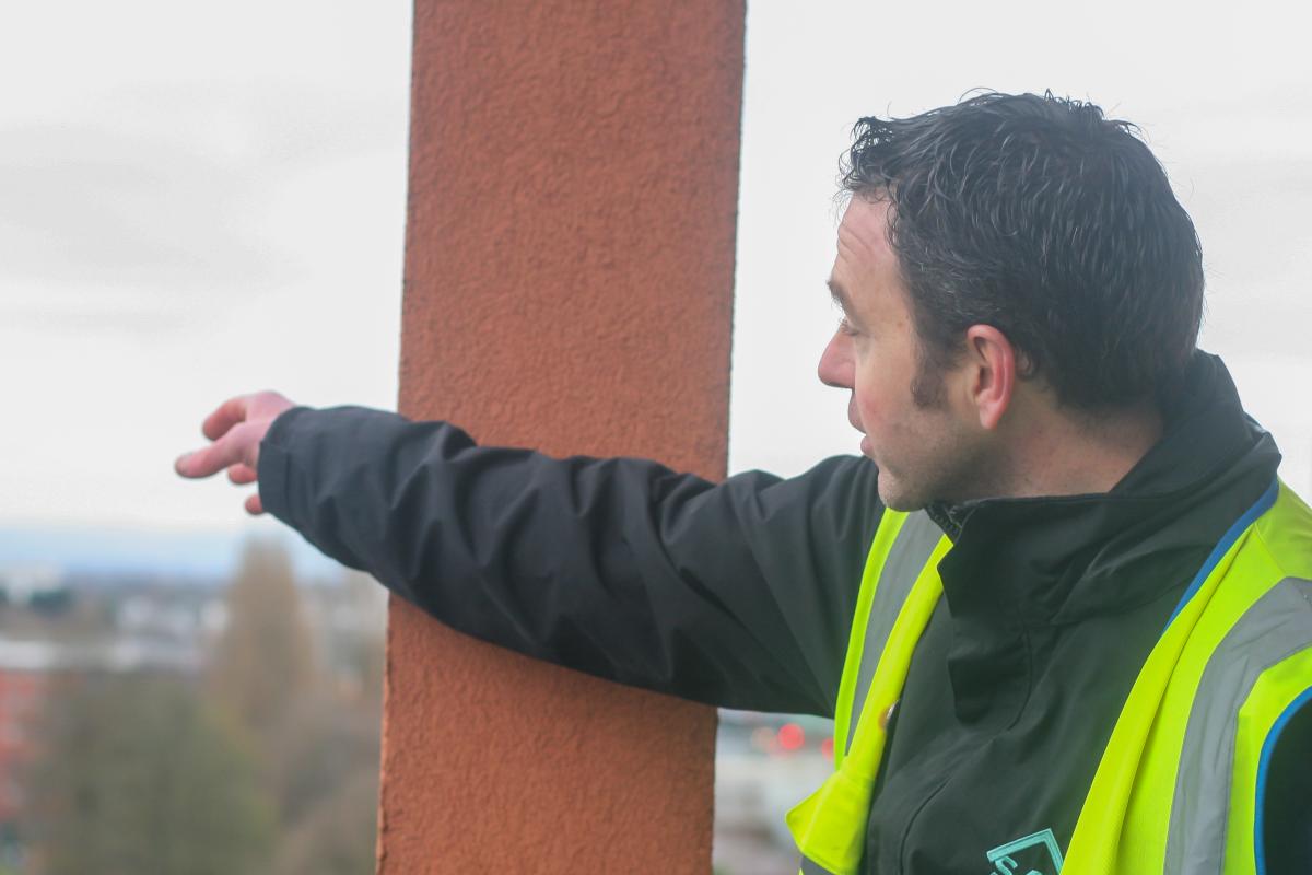 Ross Hemmings, a man with dark hair wearing a high visibility vest pointing with a red column behind him