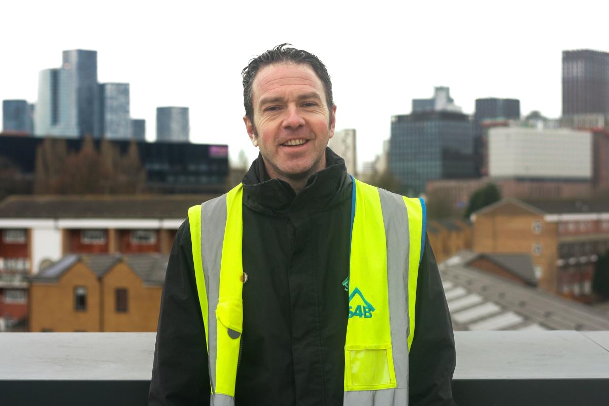 Ross Hemmings, a man with dark hair wearing a high visibility vest, standing in front of the Manchester skyline