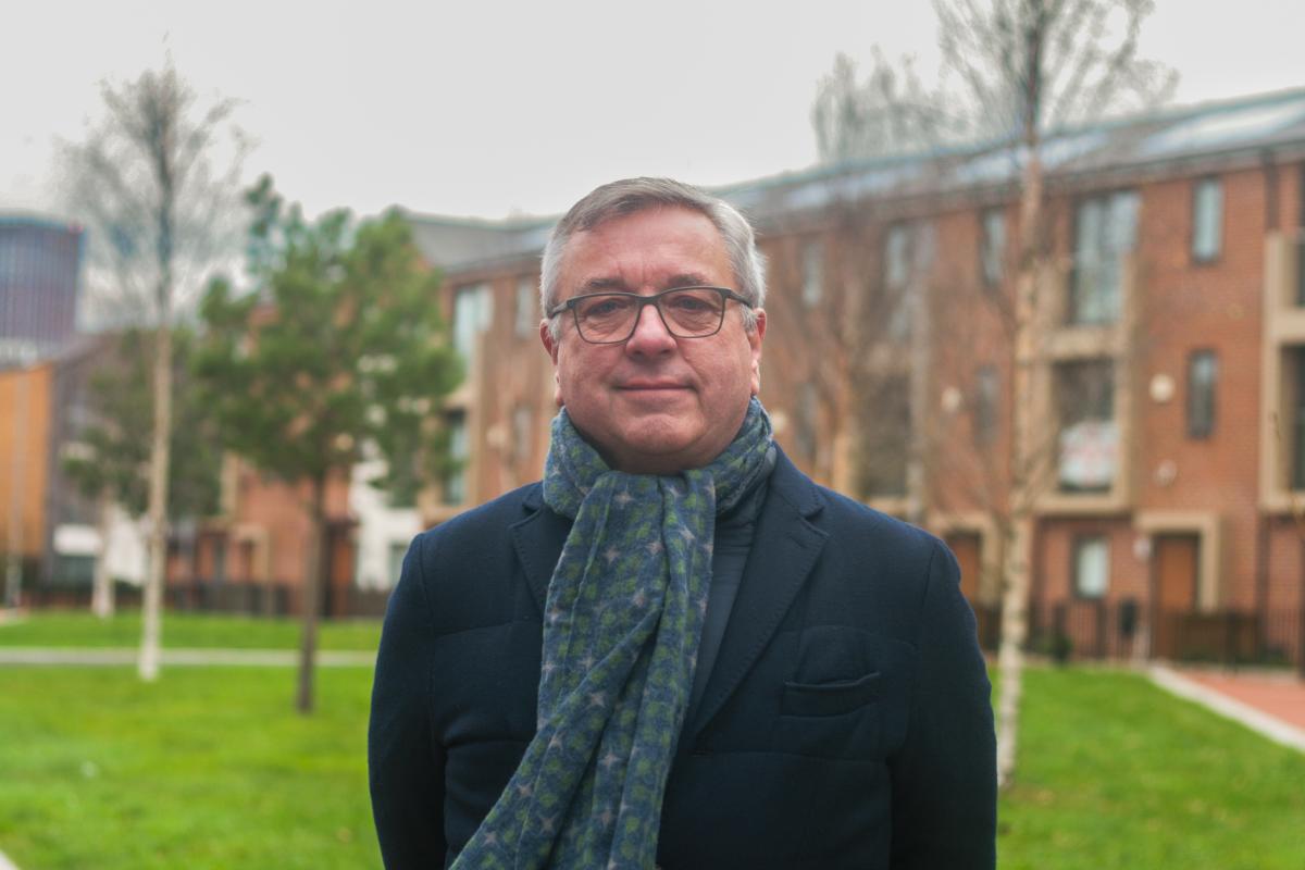 Headshot of Andy Avery, a man in a dark jacket and scarf, facing the camera in front of a green area and red brick houses.