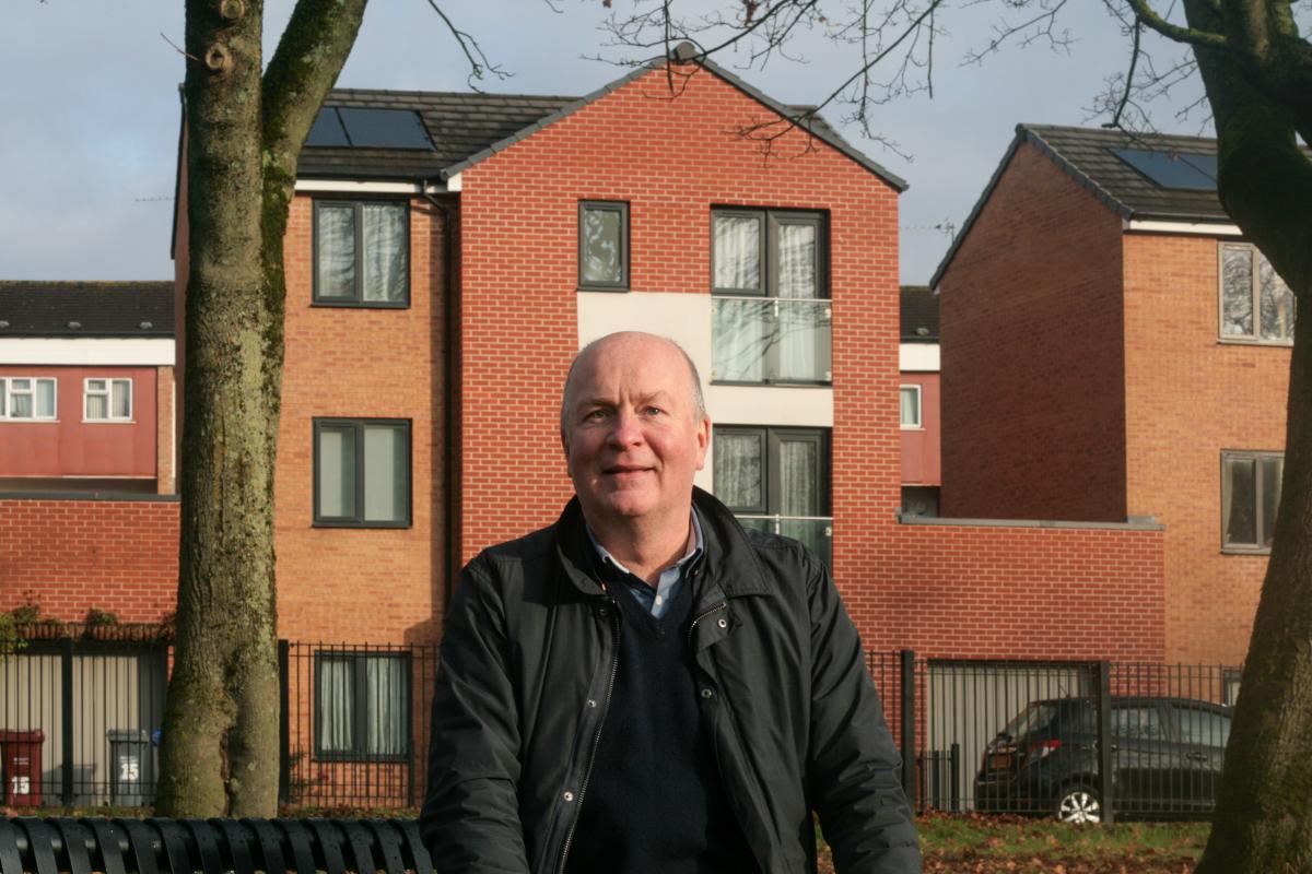 A man wearing a dark jacket sitting on a bench in front of a red brick building.