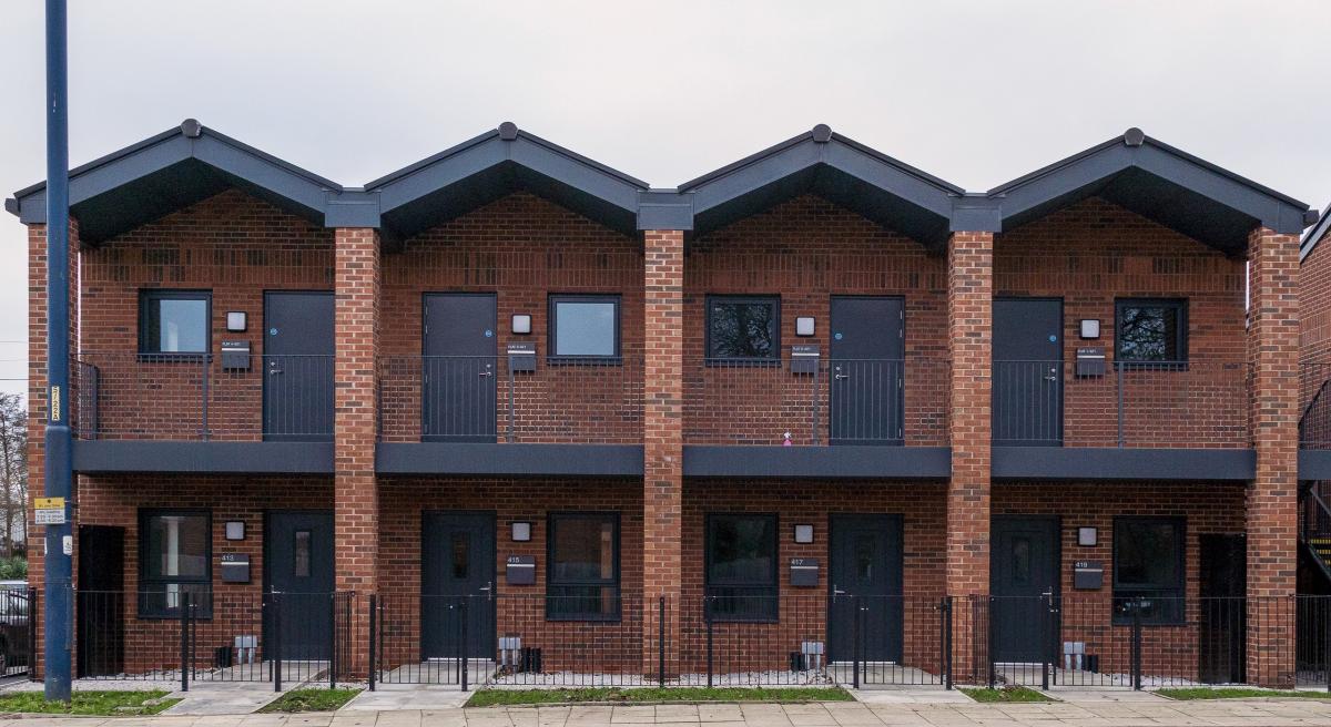 This image shows a modern row of red-brick terraced housing with gabled roofs, black-framed doors and windows, and small balconies on the upper floor. Each unit features a minimal front garden enclosed by a black metal fence, with a paved pedestrian area and street in the foreground. The design reflects contemporary urban housing, blending clean lines and traditional materials