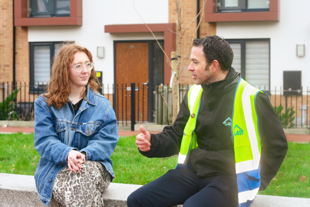 a man in a hi vis vest and woman in a denim jacket sit talking in front of some houses in Brunswick