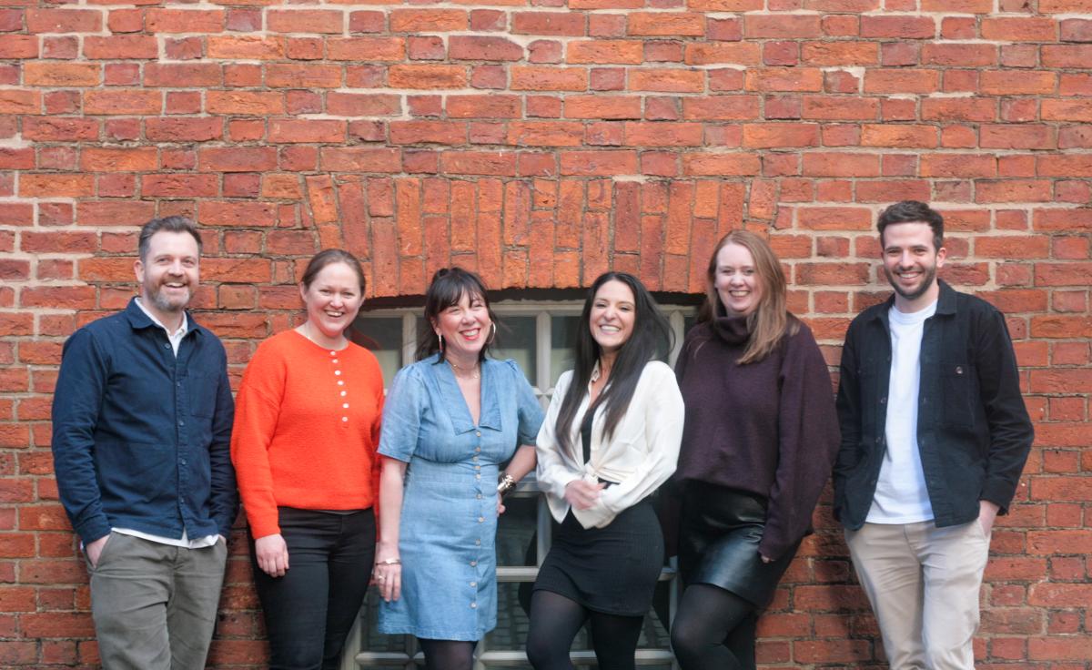 Six people - four women and two men - stood outside in front of a red brick wall