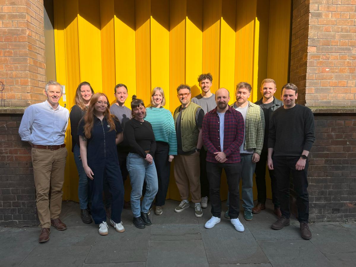 A group photo of thirteen people, outside on a street in front of a bright yellow shutter.