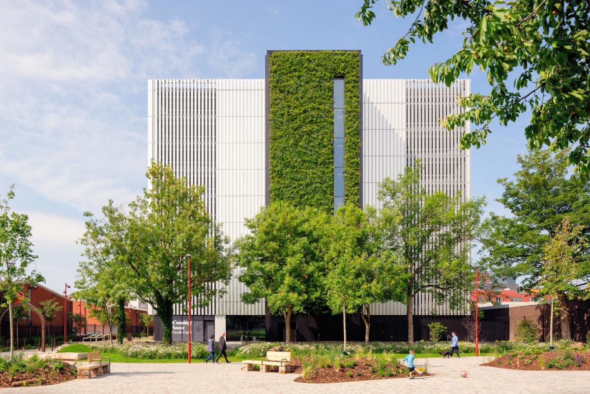Urban scene with a modern building, landscaped greenery, people walking and sitting, and a sign for a 'Coming Soon Cycle Cafe'.