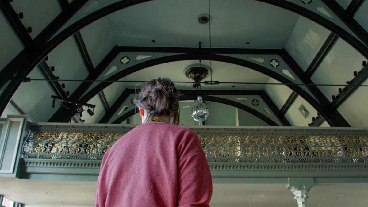 a woman in a pink jumper standing in a historic hall, looking at the ceiling.