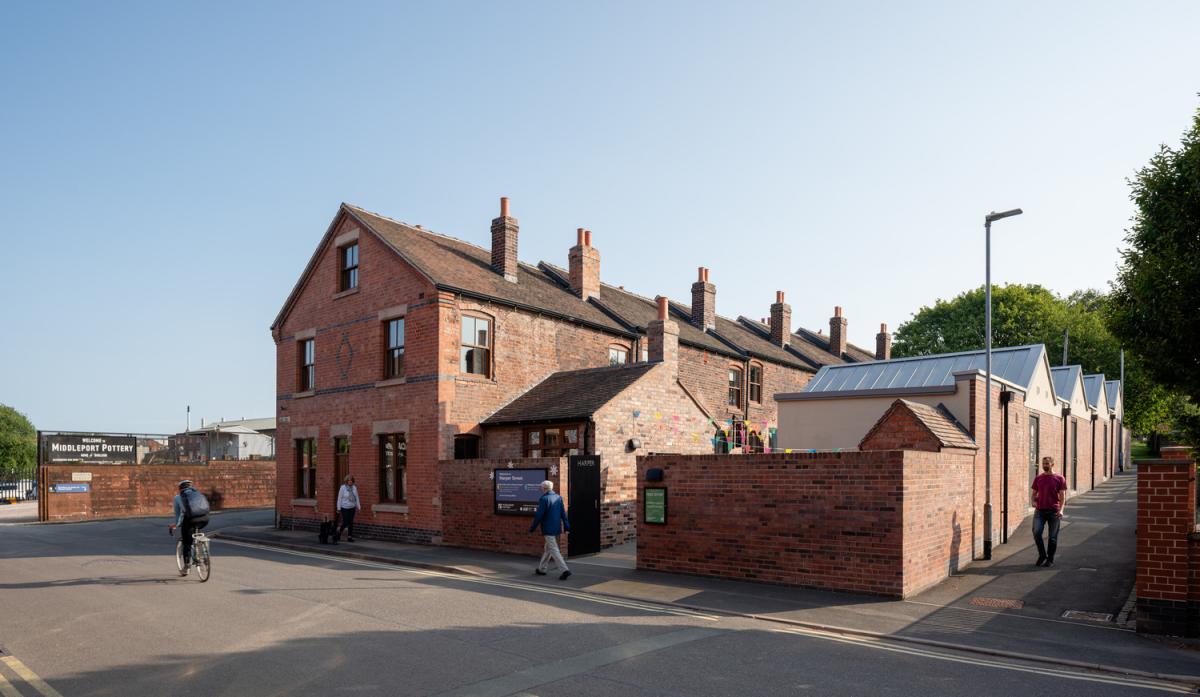 Street-level view of a corner red-brick building with pitched roof and chimneys, part of a row of terraced houses. A brick wall encloses a courtyard area. People are walking and cycling along the pavement. A signboard near the entrance reads “Middleport Pottery.”