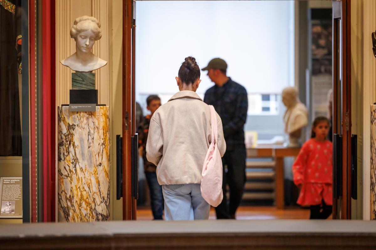 A person with a light-colored jacket and pink bag walks toward a room displaying marble busts and other sculptures. The setting includes classical columns and polished stone surfaces.