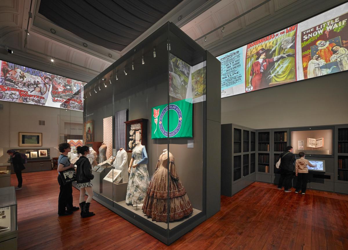 Gallery interior featuring a central glass display case with historical garments and textiles, including ornate dresses and a green banner. Visitors are viewing exhibits around the case. The room has polished wooden floors, dark shelving along the walls, and a high ceiling with projected vintage illustrations and posters near the top.
