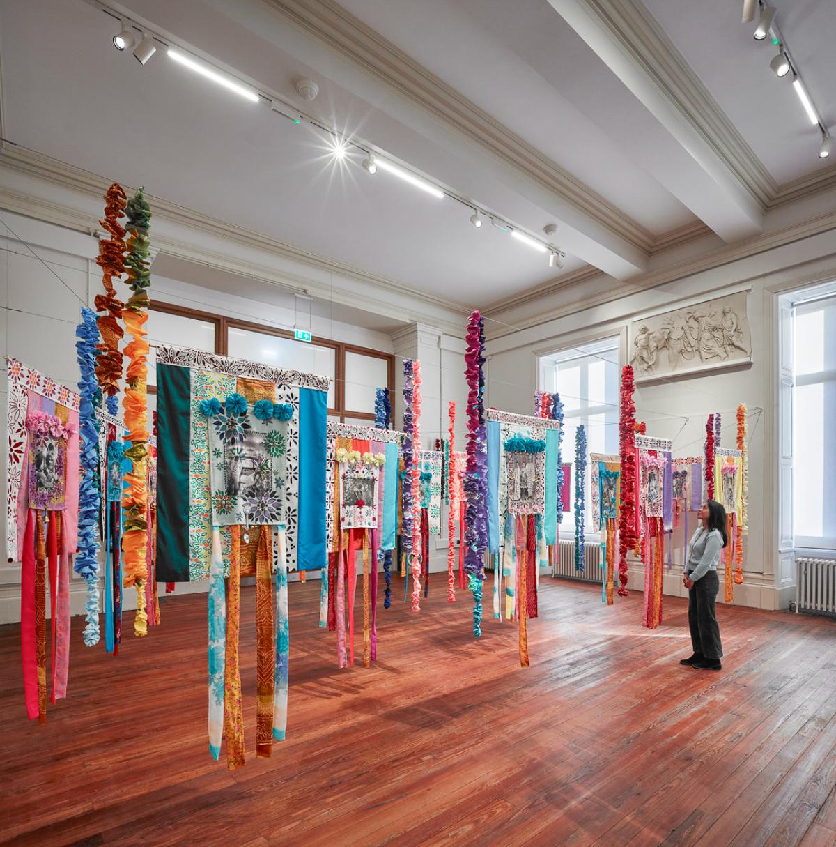 A gallery featuring vibrant hanging textile pieces with layered patterns and ribbons, arranged in rows across a polished wooden floor.