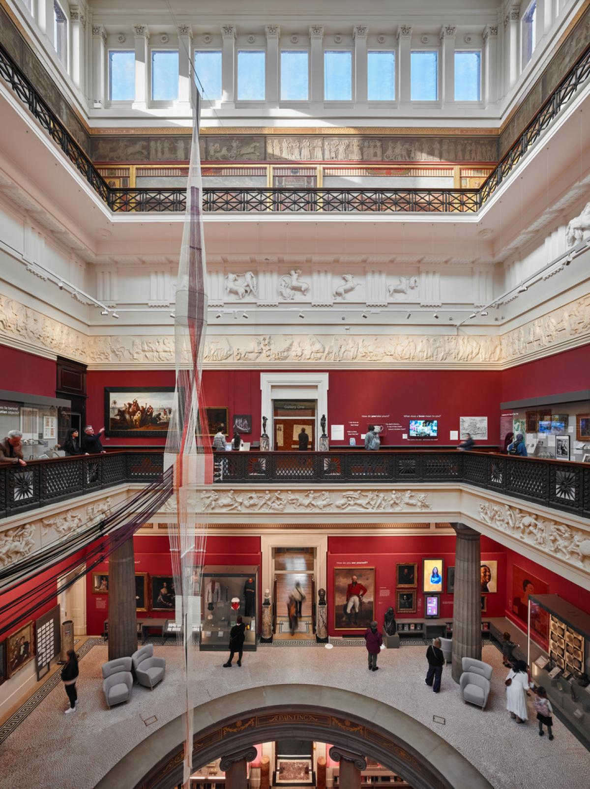 Inside a museum setting, with balconies and a central atrium, red walls and people on the different levels.