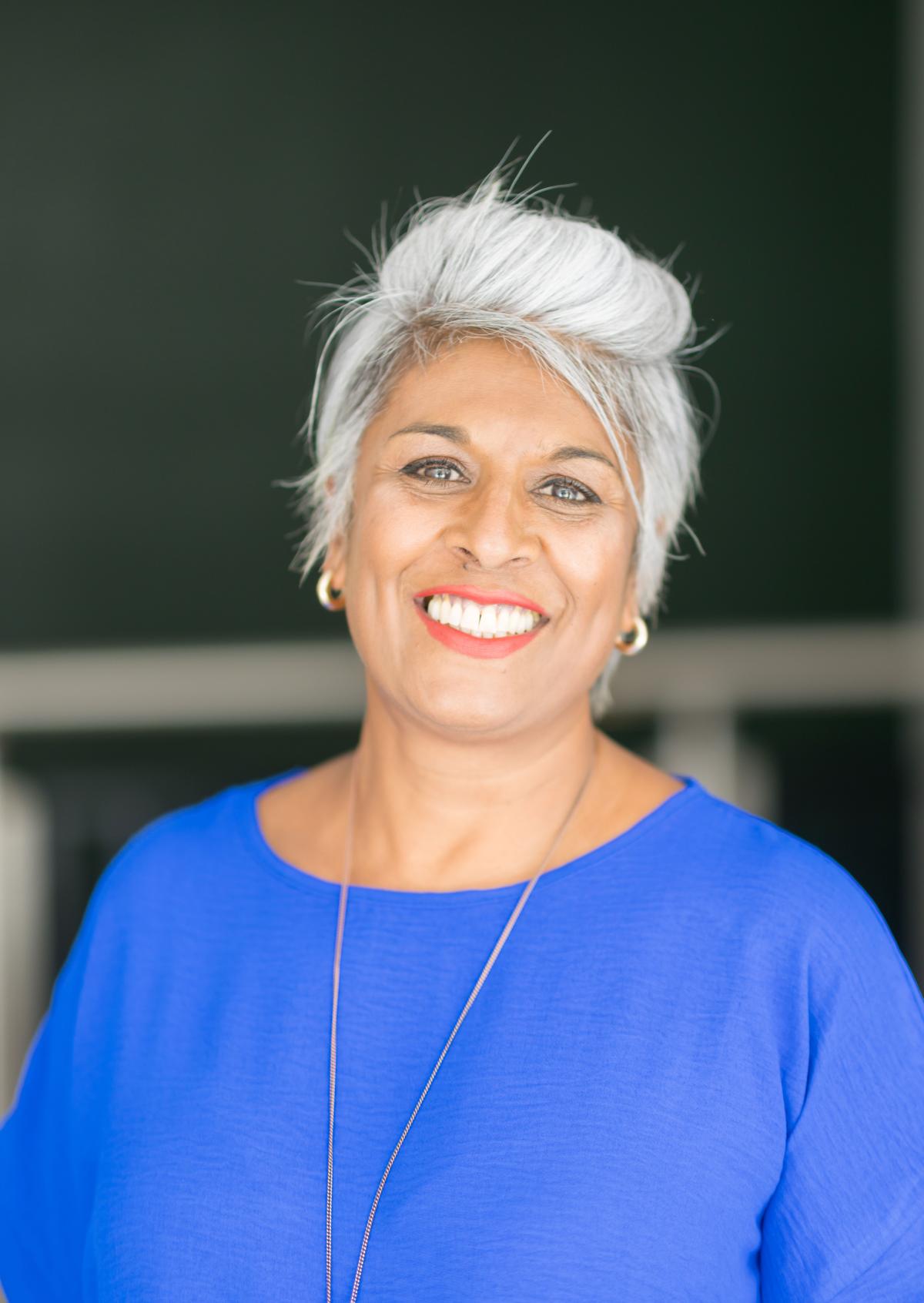 Woman with brown skin wearing a blue top, short grey hair and smiling.