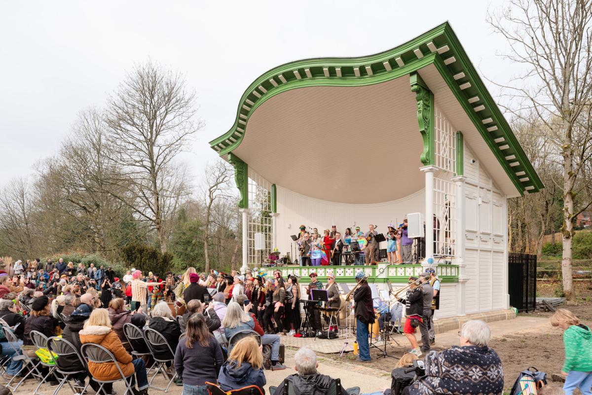 Community brass band and singers in colorful costumes perform on a green-and-white Victorian-style bandstand while a large seated audience watches on.