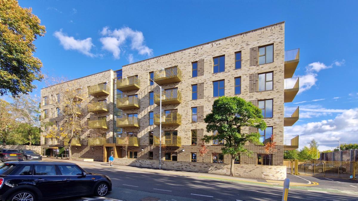 A light brick new apartment block with golden coloured balconies, situation street side with trees around.