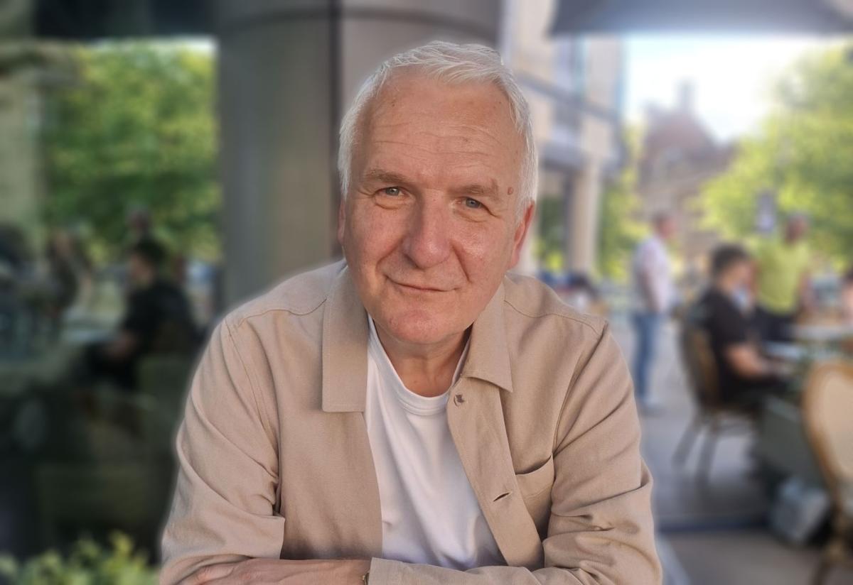 A man with grey hair, wearing a beige open first and white t-shirt underneath sat outside smiling at the camera