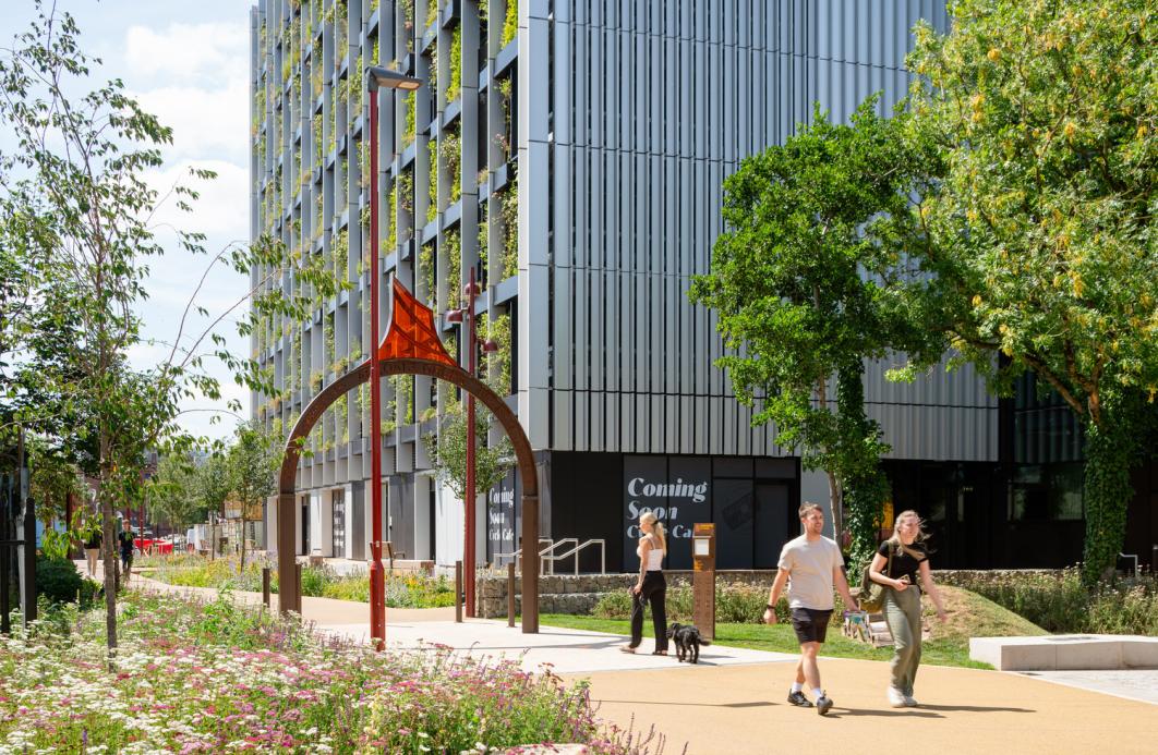 Modern building with integrated greenery, a flower-lined path, two people walking—one with a dog—and an archway sculpture; 'Coming Soon' sign visible.