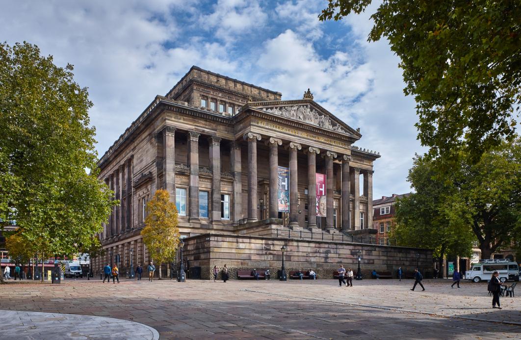 A neoclassical building with tall stone columns and ornate pediment, surrounded by trees and a paved square where people are walking.