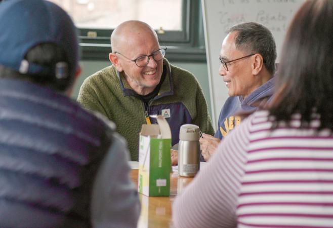 two men are sat in a group around a table smiling and having a conversation. They are learning english as a second language at stretford public hall
