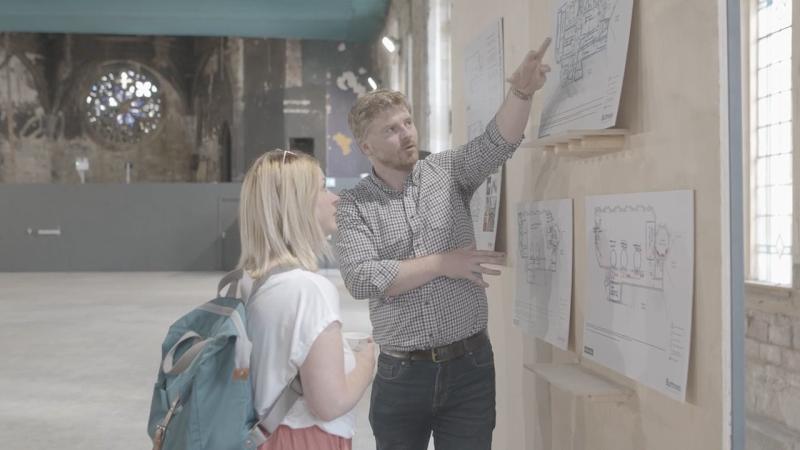 Internal photograph of a man showing a woman an architectural drawing on a noticeboard on a wall