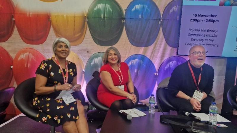 Internal image of three people sitting behind a coffee table. Infront of them are their speaker's notes. Behind is a large colourful screen with an Amazon logo. Chithra Marsh is sitting on the left. 