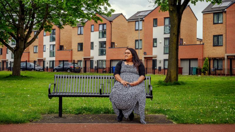 a lady sits on a bench in a park. A row of brick houses sit behind.