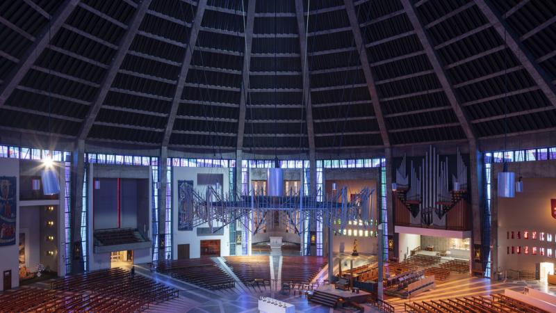 Internal photograph of Liverpool Metropolitan Cathedral. It's a circular building with rich colours of red, purple and yellow. Seating is laid out in a circular fan-style with the pulpit in the middle of the circle.  