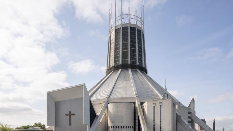 External photograph of a teepee shaped church made out of grey concrete. On the top half of the building there is a circular crown lantern made from coloured glass and finished with tall spires. 