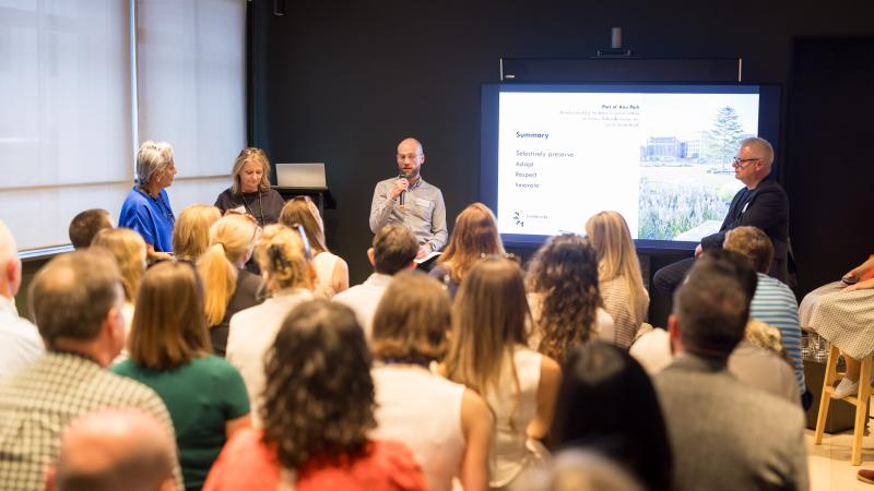 Audience of people facing four panellists and a presentation screen in a room.